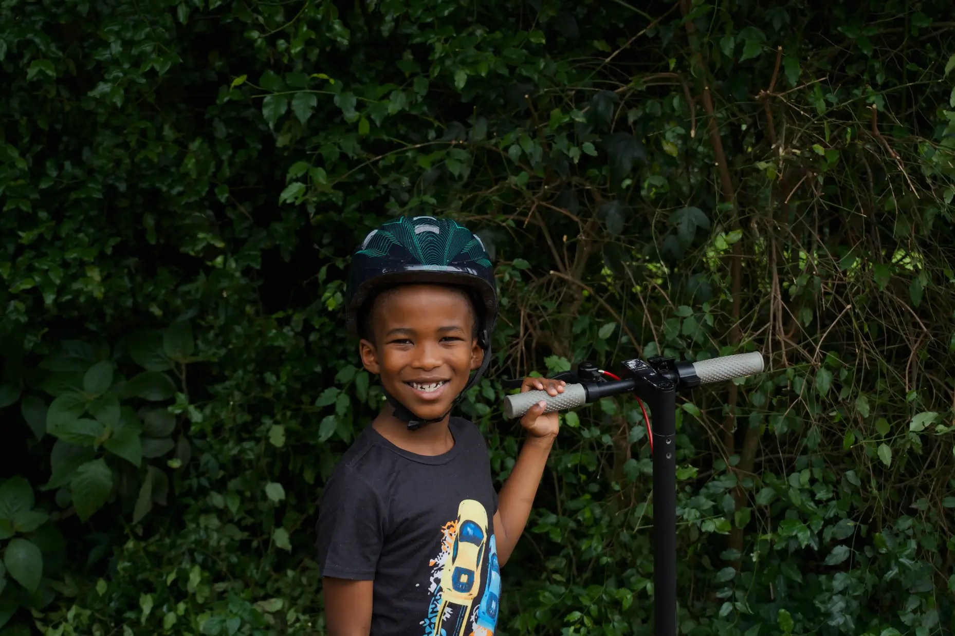 A medium shot of a boy smiling in front of a tall shrub, with his electric scooter.